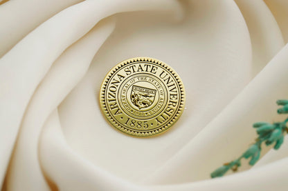 A gold-colored brooch with an engraved university seal placed on a white fabric with a floral pattern.