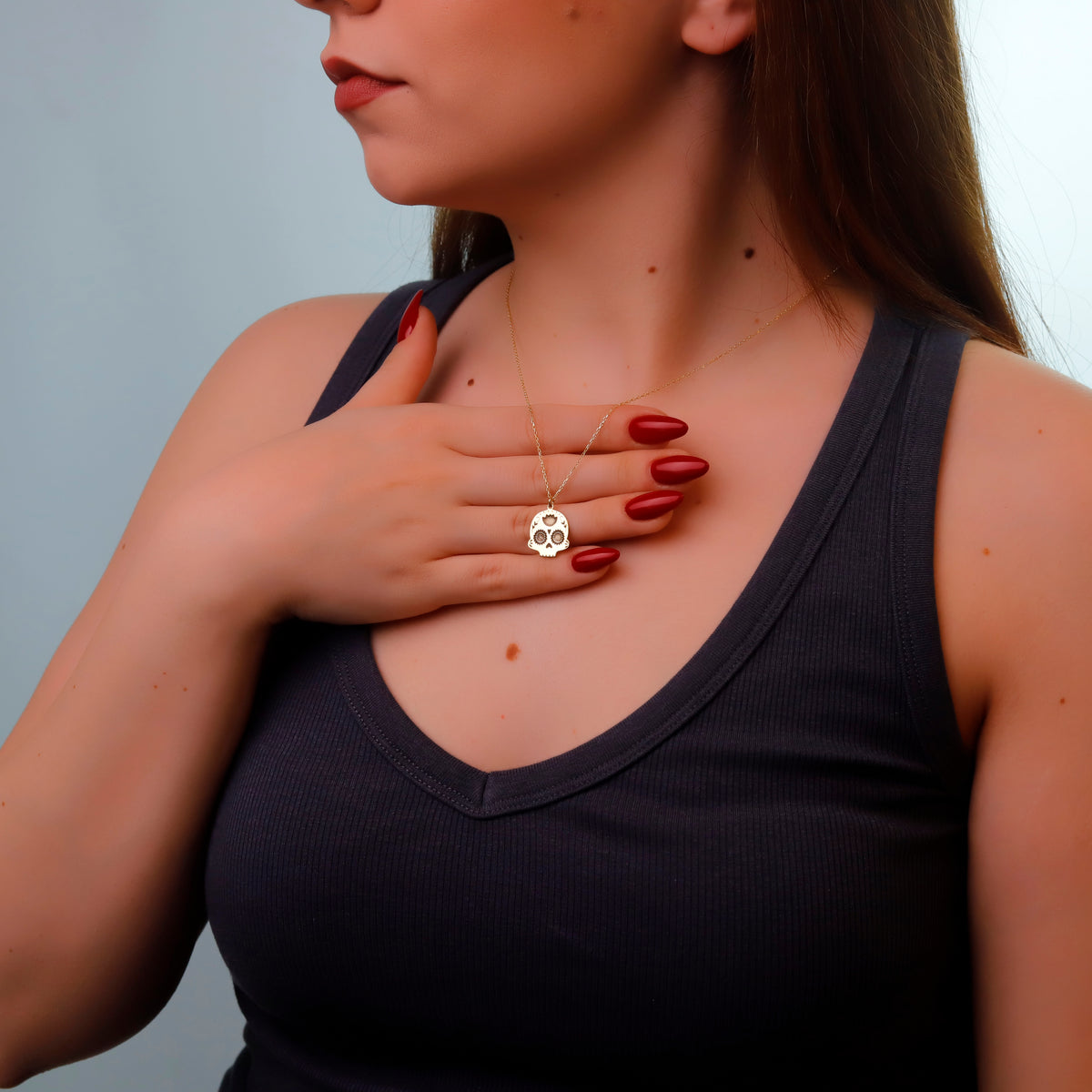 Woman wearing a black tank top and gold necklace with a skull pendant against a gray background