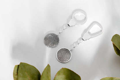 A sterling silver keychain with a fingerprint-like pattern on it, held between fingers with a neutral background.