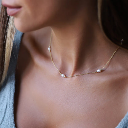 A close-up image of a woman wearing a gold choker necklace with freshwater pearls, over a light blue spaghetti strap top.
