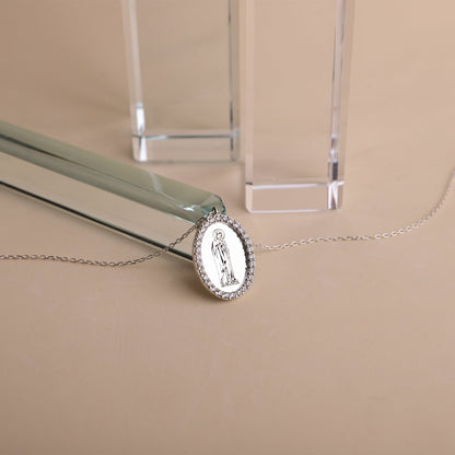 A silver necklace featuring a round pendant with an image of the Virgin Mary, encapsulated by cubic zirconia crystals, displayed on a flat surface.