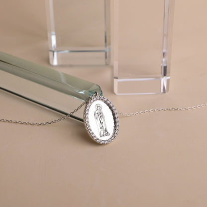 A silver necklace featuring a round pendant with an image of the Virgin Mary, encapsulated by cubic zirconia crystals, displayed on a flat surface.