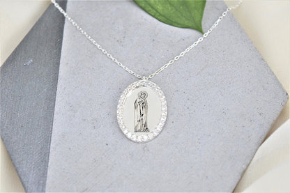 A silver necklace featuring a round pendant with an image of the Virgin Mary, encapsulated by cubic zirconia crystals, displayed on a flat surface.