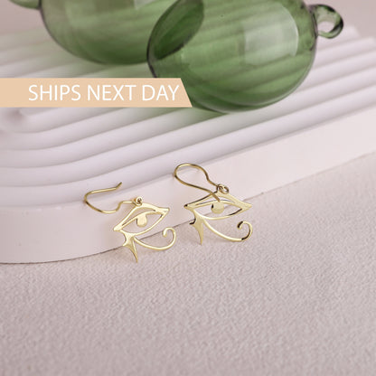 Gold eye-shaped earrings on a light surface with green glass objects in the background.