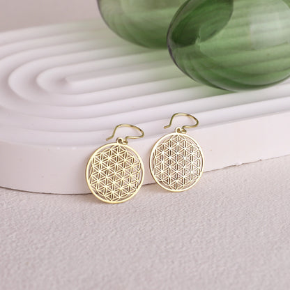 A pair of gold earrings featuring the Flower of Life pattern, displayed on a white stand with a green glass object in the background.