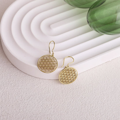 A pair of gold earrings featuring the Flower of Life pattern, displayed on a white stand with a green glass object in the background.