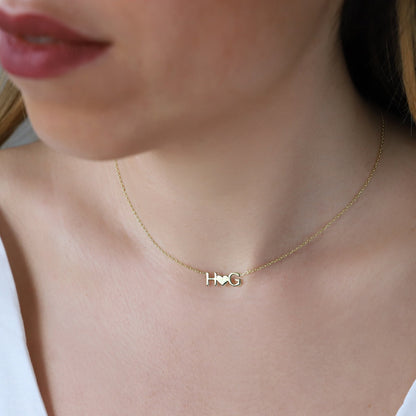 A woman wearing a gold-colored necklace with a heart-shaped pendant that has an initial letter 'H' on it.
