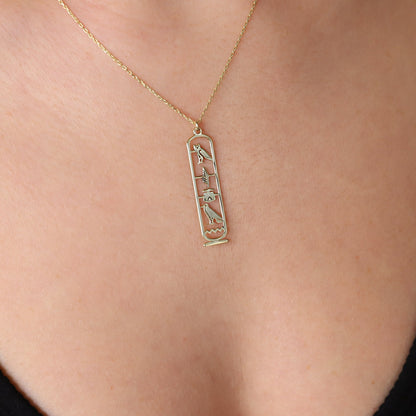 A close-up image of a person wearing a gold-colored Egypt cartouche necklace with a hieroglyph pendant.