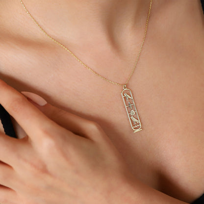 A close-up image of a person wearing a gold-colored Egypt cartouche necklace with a hieroglyph pendant.