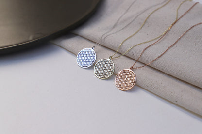 A close-up image of a minimalist gold pendant necklace with the Flower of Life pattern, being held by a person wearing a white top.