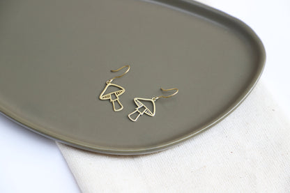 A person wearing gold mushroom dangle earrings against a blurred background.