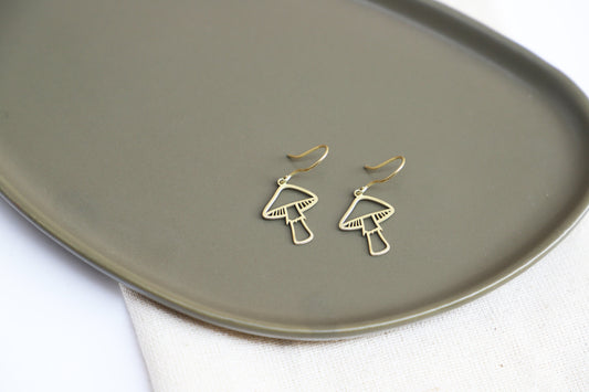 A person wearing gold mushroom dangle earrings against a blurred background.