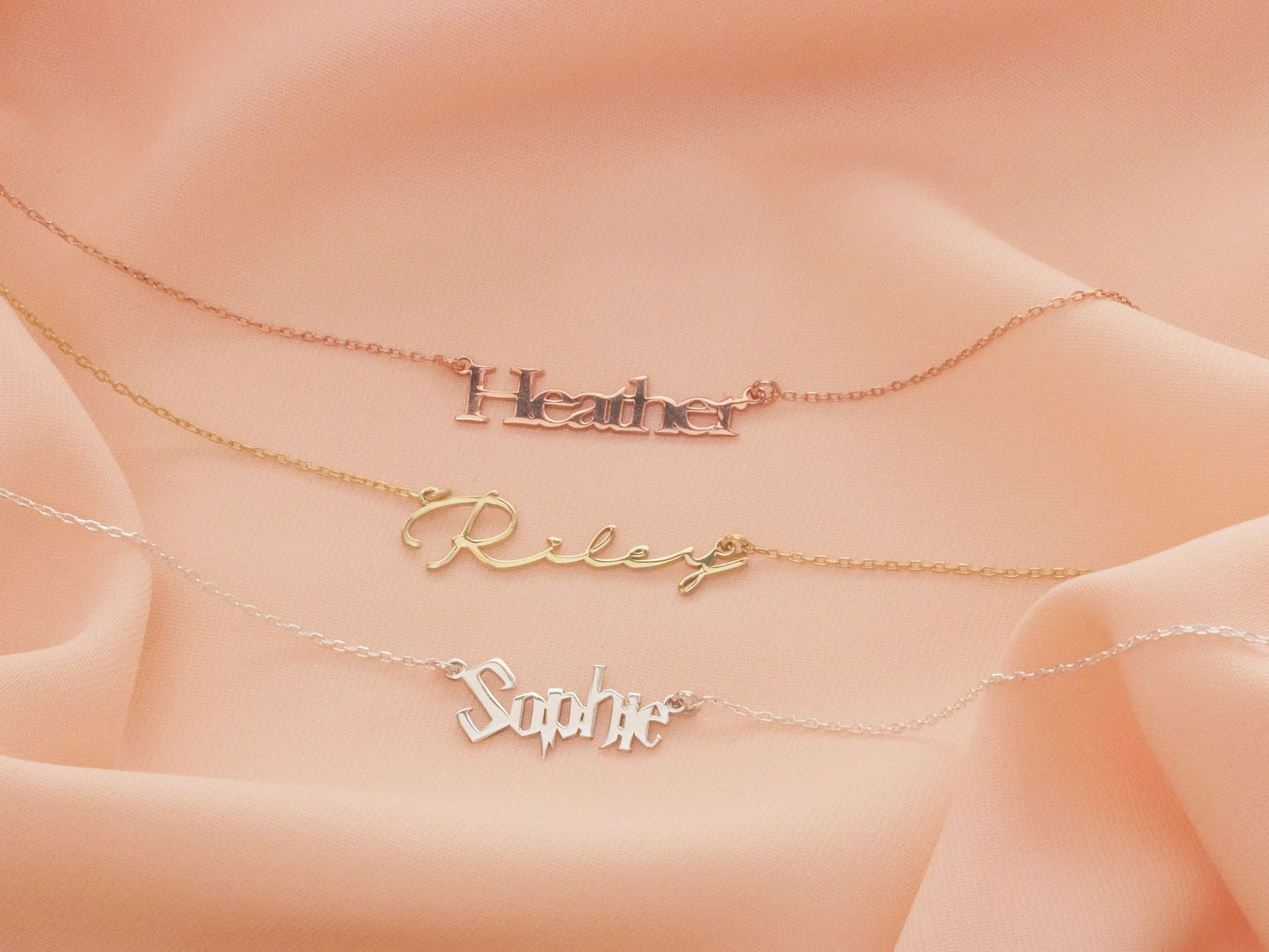 Three gold name necklaces on a soft pink fabric background