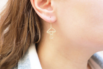 A person wearing gold mushroom dangle earrings against a blurred background.