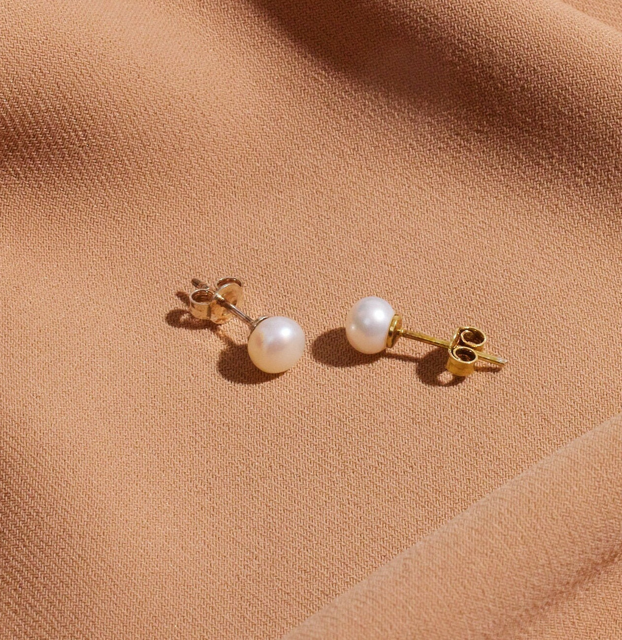 A pair of gold-colored pearl stud earrings placed on a brown surface.