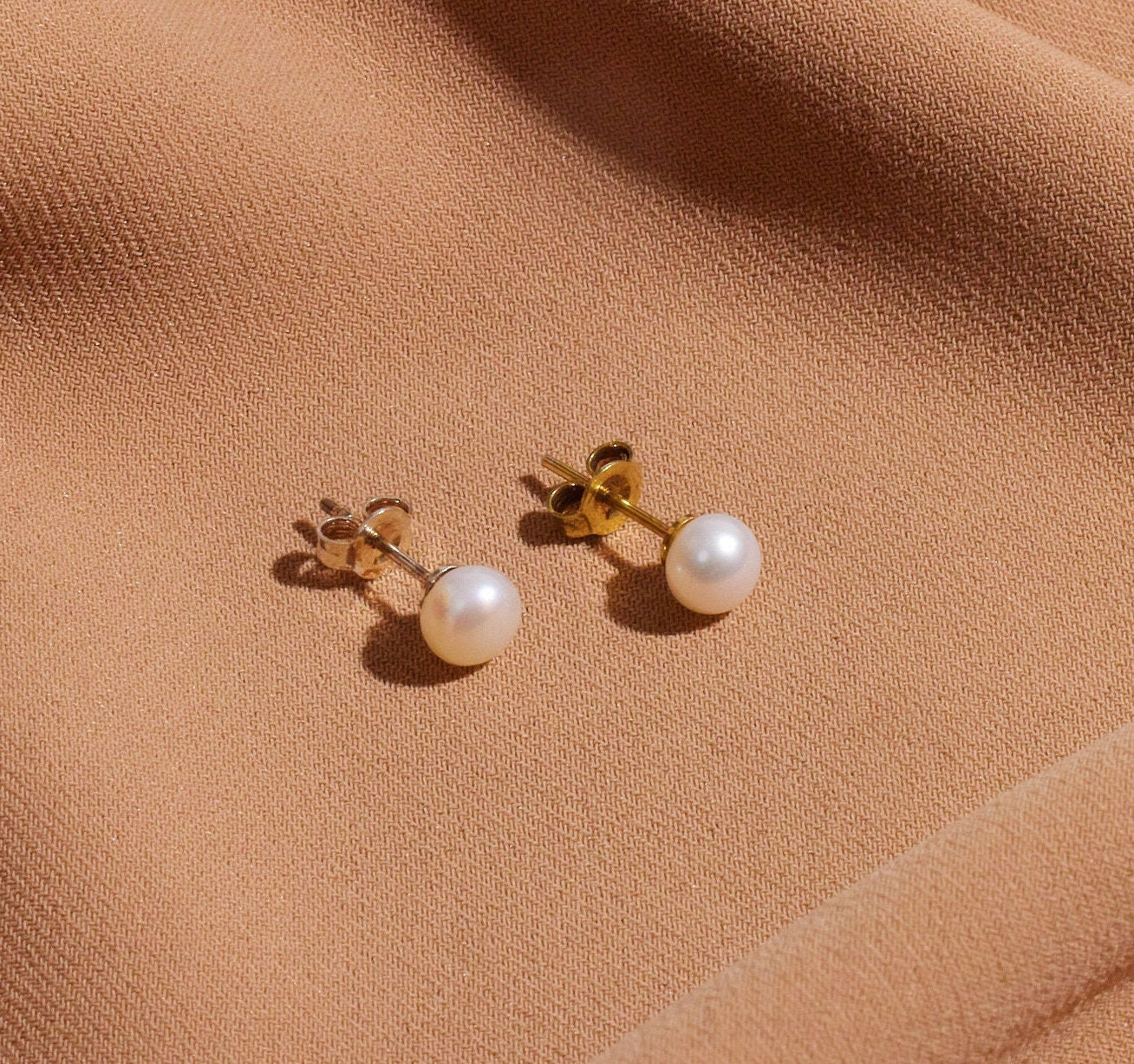 A pair of gold-colored pearl stud earrings placed on a brown surface.