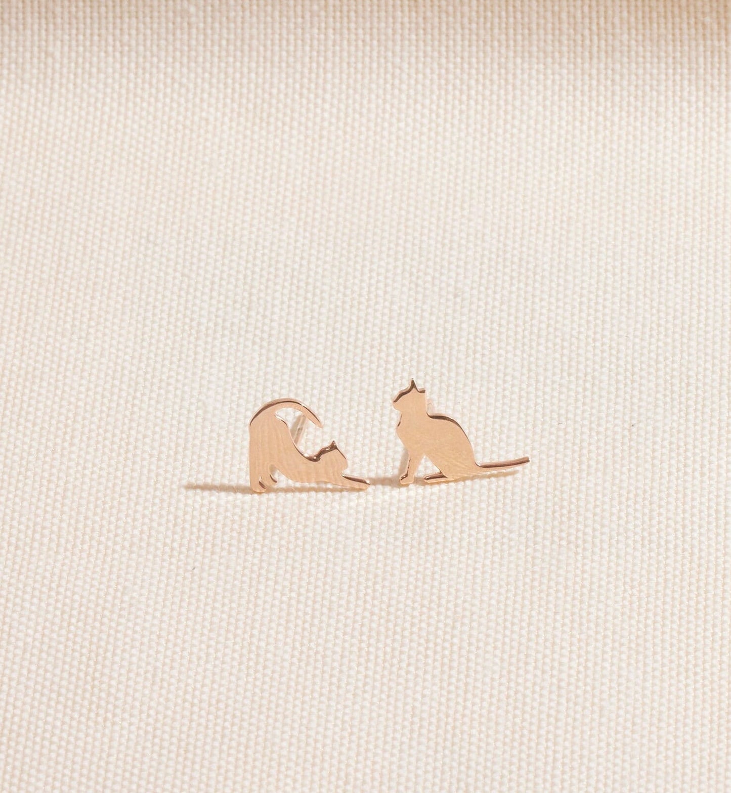 Rose gold cat-shaped earrings on a beige fabric background