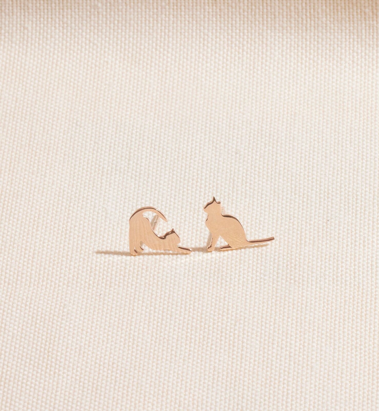 Rose gold cat-shaped earrings on a beige fabric background