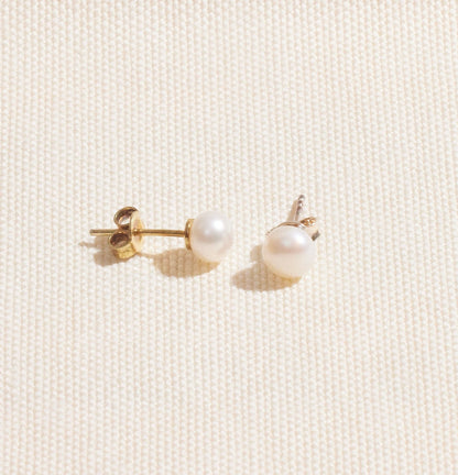 A pair of gold-colored pearl stud earrings placed on a white surface.