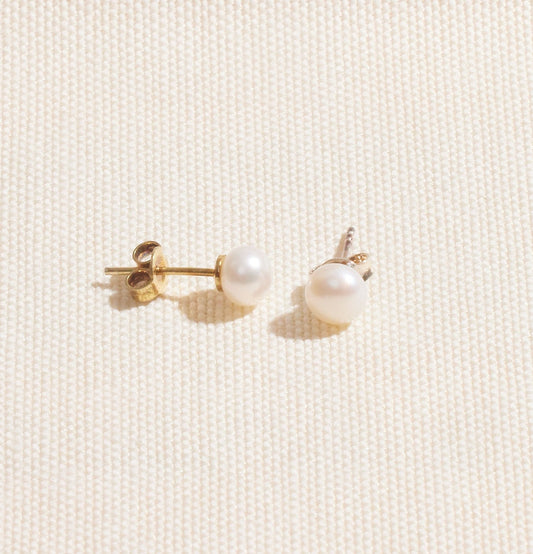 A pair of gold-colored pearl stud earrings placed on a white surface.