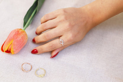 A hand wearing a gold heart knot ring, with two additional rings on the table beside a pink tulip.