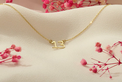 A close-up image of a dainty necklace with the number '13' on it, showcasing the necklace's delicate design and the chain's texture.