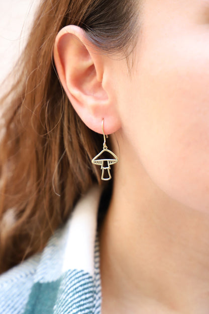 A person wearing gold mushroom dangle earrings against a blurred background.