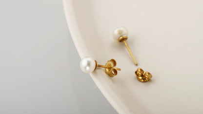 A pair of gold-colored pearl stud earrings placed on a white surface.