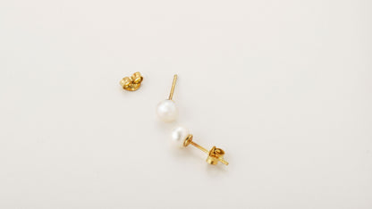 A pair of gold-colored pearl stud earrings placed on a white surface.
