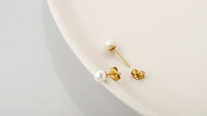 A pair of gold-colored pearl stud earrings placed on a white surface.