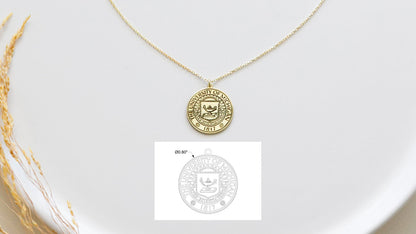 A gold necklace with a circular pendant featuring a custom logo, displayed on a white plate with a small paper insert showing the logo design.