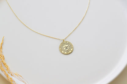 A gold necklace with a circular pendant featuring a custom logo, displayed on a white plate with a small paper insert showing the logo design.