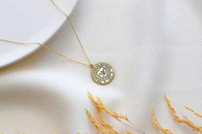 A gold necklace with a circular pendant featuring a custom logo, displayed on a white plate with a small paper insert showing the logo design.