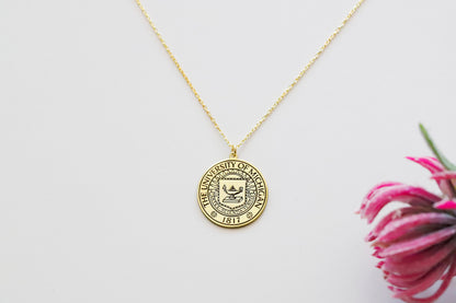 A gold necklace with a circular pendant featuring a custom logo, displayed on a white plate with a small paper insert showing the logo design.