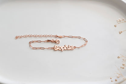 A personalized name bracelet with link chain and add-on birthstone, displayed on a white plate with dried flowers around it.