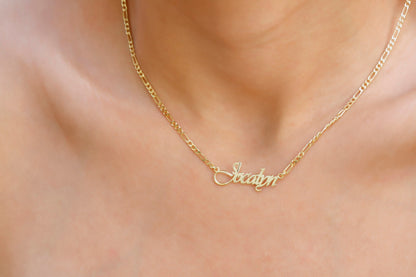 A gold-colored Figaro chain necklace with a customizable cursive name pendant.