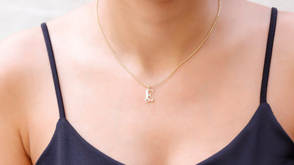 A person wearing a black tank top showcasing a white gold ball chain necklace with a single initial 'E' and a tiny heart pendant.