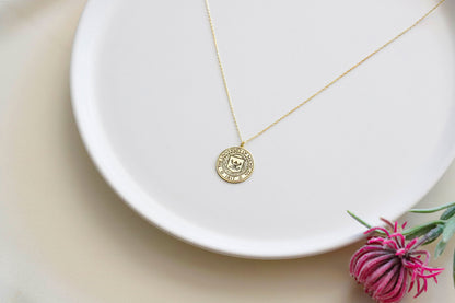 A gold necklace with a circular pendant featuring a custom logo, displayed on a white plate with a small paper insert showing the logo design.