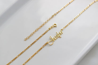 A gold-colored Figaro chain necklace with a customizable cursive name pendant.
