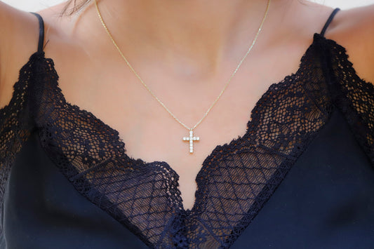 A close-up image of a person wearing a 14K gold diamond cross necklace with a delicate chain, displayed against a lacy black background.