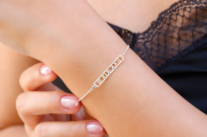 A sterling silver bar bracelet with Roman numerals engraved on it, worn on a person's wrist.