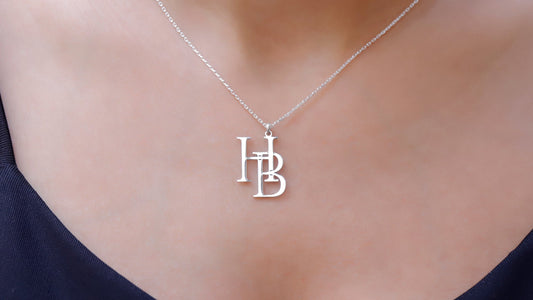 Two interlocking custom design letter necklaces in gold color, with the letters 'K' and 'B' respectively, displayed on a white fabric surface.