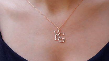 Two interlocking custom design letter necklaces in gold color, with the letters 'K' and 'B' respectively, displayed on a white fabric surface.