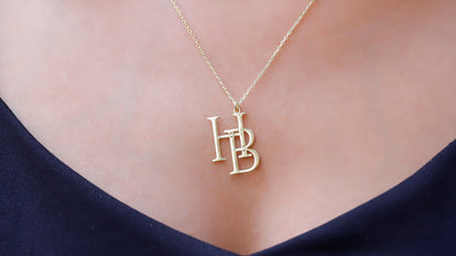 Two interlocking custom design letter necklaces in gold color, with the letters 'K' and 'B' respectively, displayed on a white fabric surface.