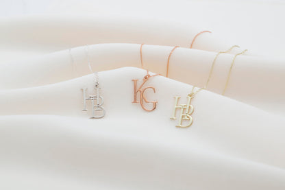 Two interlocking custom design letter necklaces in gold color, with the letters 'K' and 'B' respectively, displayed on a white fabric surface.
