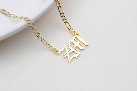 A close-up of a person wearing a gold-filled necklace with a customizable charm of the letters 'ZM' in a Gothic font. A product by SilverGoldandRose.com.