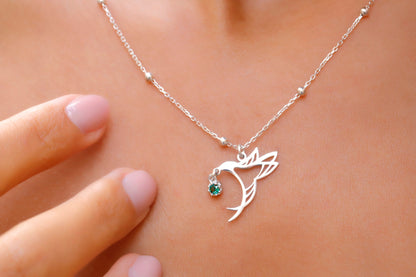 A close-up image of a woman's neck wearing a silver-colored necklace with a bird charm and beaded ball chain.