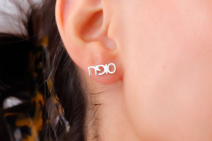 A close-up image of a woman's ear wearing a small gold-colored stud earring with a personalized Hebrew name.