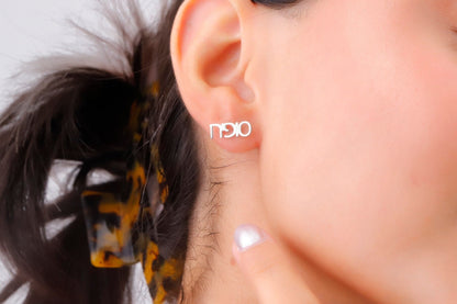 A close-up image of a woman's ear wearing a small gold-colored stud earring with a personalized Hebrew name.