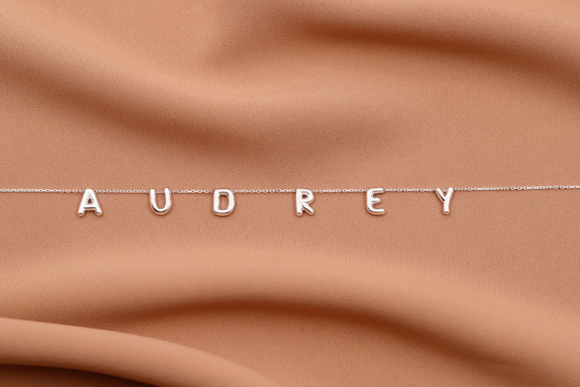 Silver necklace with 'AUDREY' letters on a brown fabric background